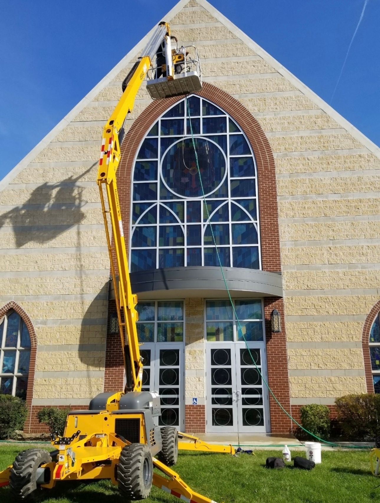 A worker in a yellow boom lift cleans the large arched windows of a tan brick church on a sunny day.