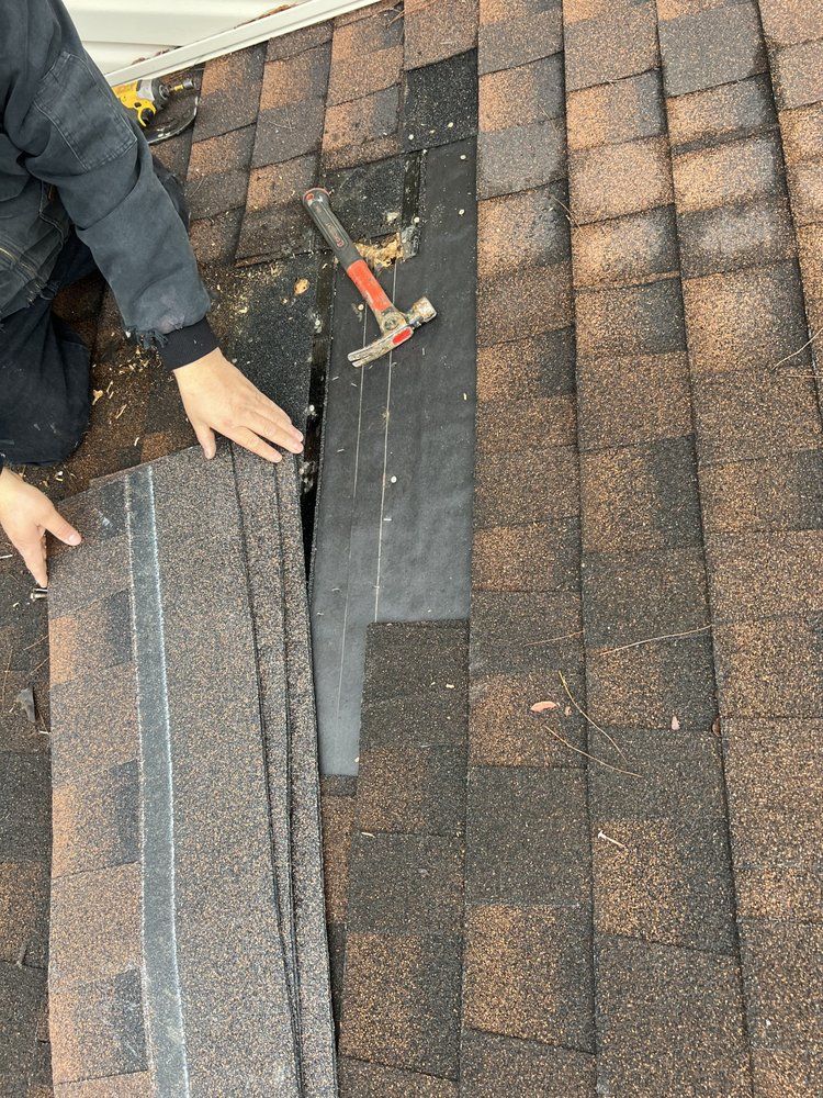 Person on a roof repairing shingles. They hold new brown shingles and a hammer rests on the exposed black underlayment.