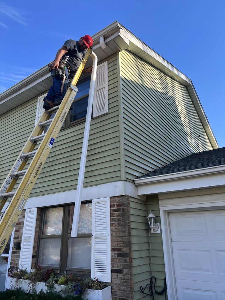 Person on a ladder installing a white gutter on a light green house with white shutters. Blue sky in the background.