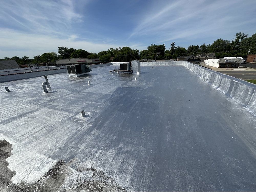 Flat commercial roof coated with a reflective silver substance under a bright blue sky. Several vents and pipes protrude from the surface.