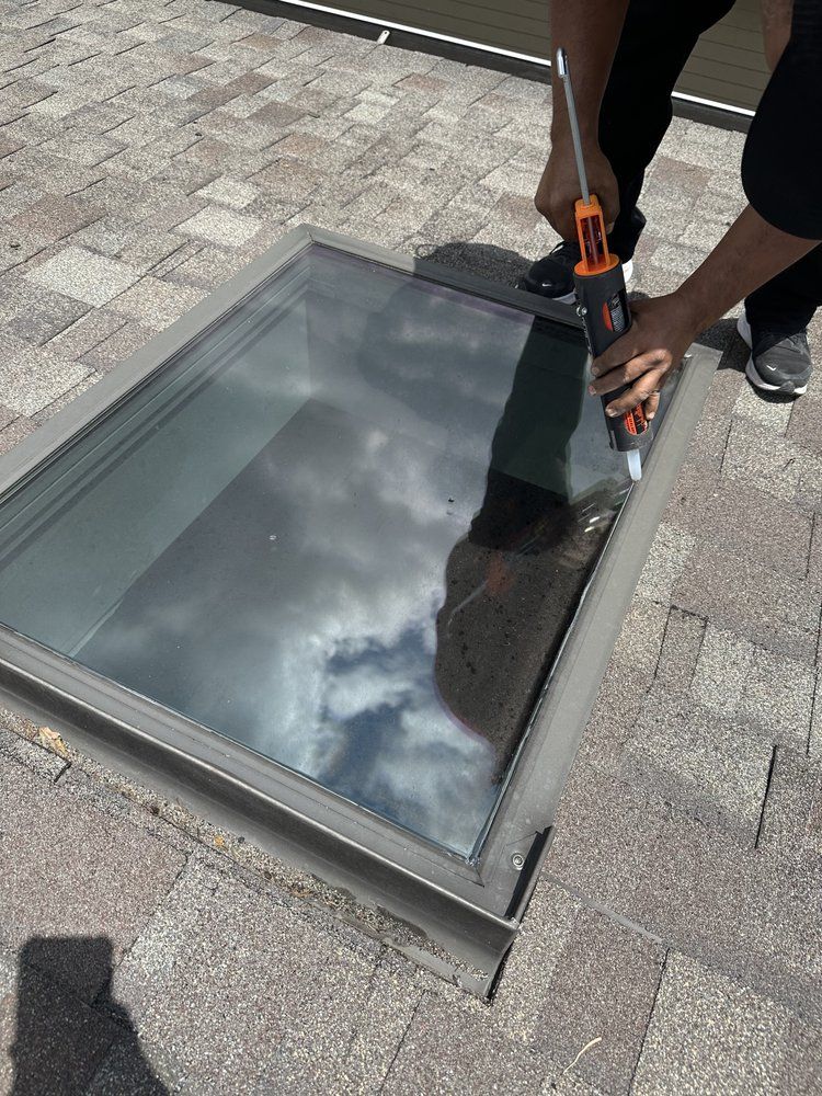 Person applying sealant around a skylight on a shingled roof. The sky is reflected in the glass.