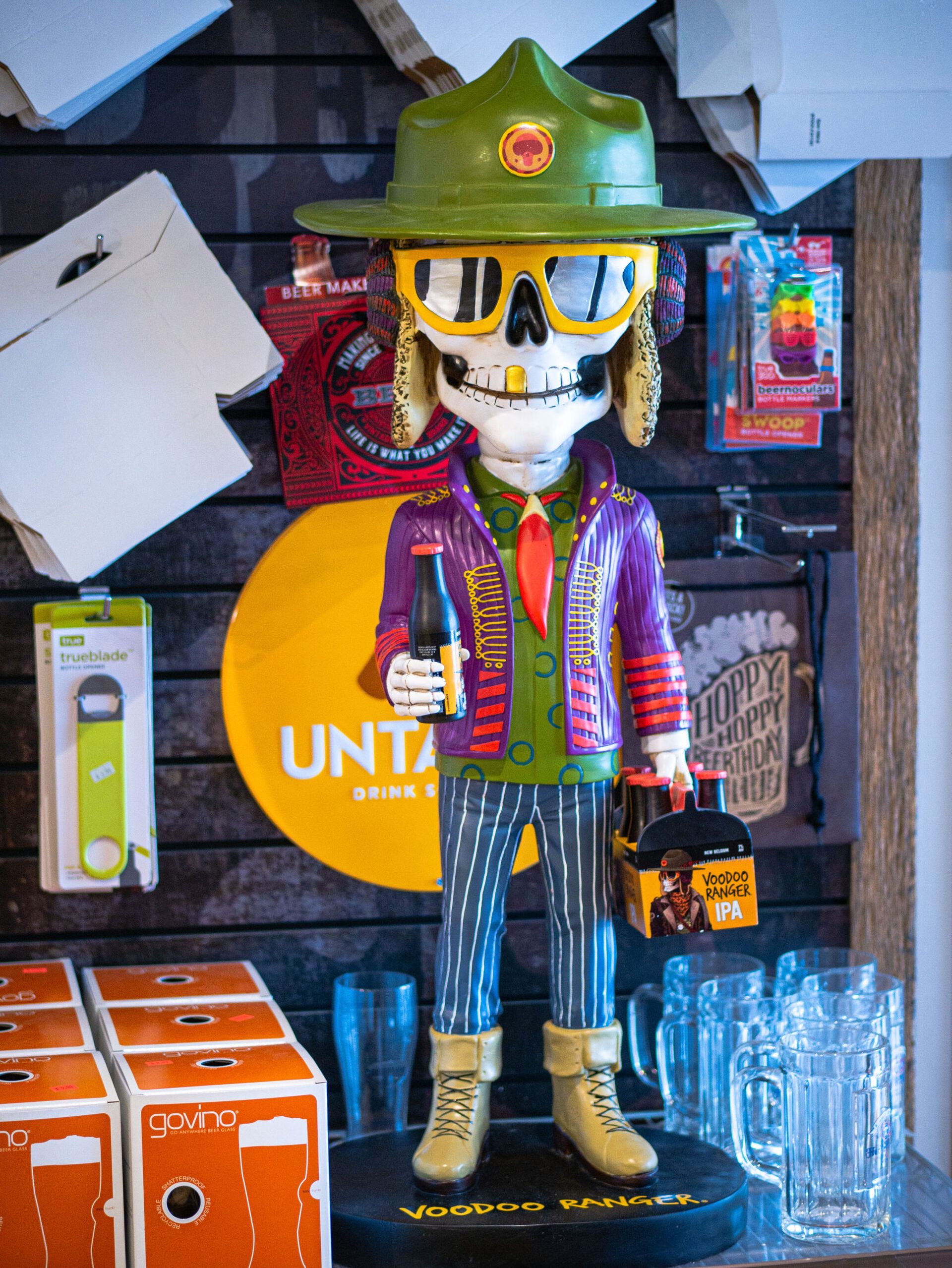 Skeleton figure in green hat and yellow sunglasses in a shop display, with colorful bottles and glassware.