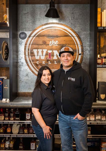 Two people standing in a bar with a large circular wall logo and shelves of bottles behind them.