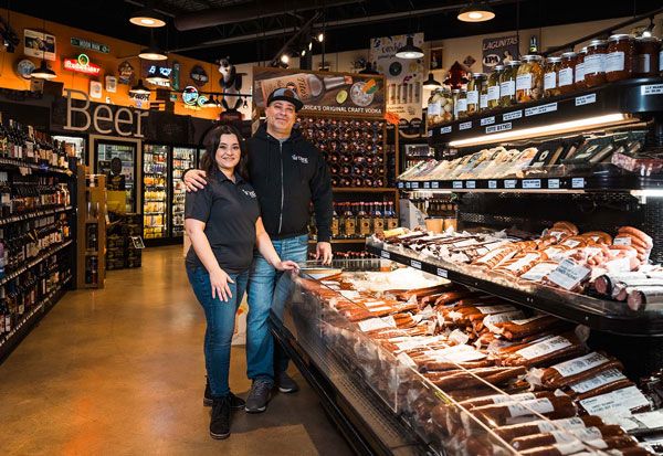 Two people standing in a grocery store deli aisle beside a refrigerated display of meats and cheeses.
