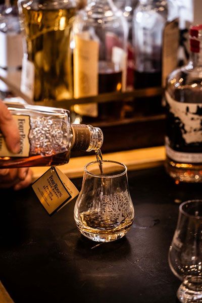 Whiskey being poured into a tasting glass at a bar, with bottles in the background.