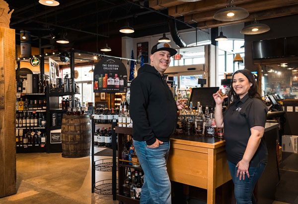 Two people standing in a liquor store aisle, smiling beside a wooden counter.