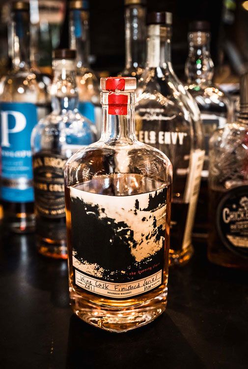 Clear liquor bottle with red cap in front of assorted whiskey bottles on a bar shelf