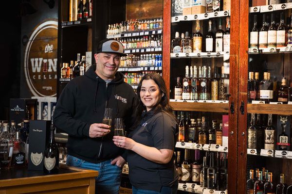 Two people holding wine glasses in a wine shop with shelves of bottles behind them.