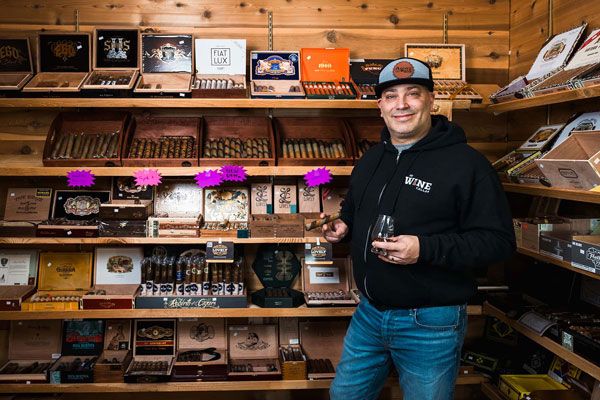 Person standing in a cigar shop, smiling beside shelves of boxed cigars and tobacco products.
