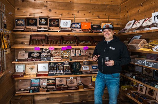 Man standing in a wooden shop beside shelves of packaged coffee products, holding a drink and smiling.