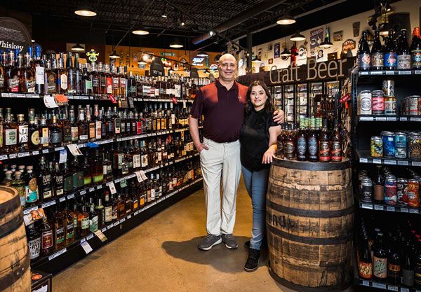 Two people standing in a liquor store aisle beside a wooden barrel display