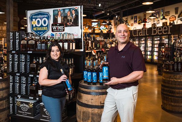 Two people standing in a craft beer store, smiling beside beer barrels and shelves of bottles.