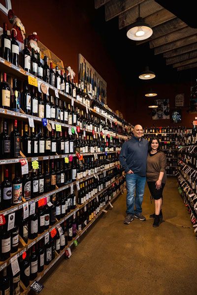 Two people standing in a narrow wine shop aisle lined with bottles and shelves
