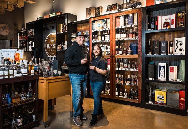 Two people standing in a liquor store aisle beside shelves of bottles and wine display cases