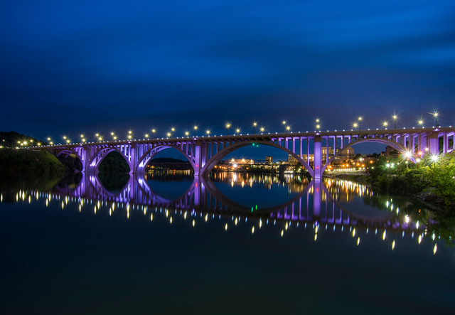 Night view of Knoxville river bank a purple-lit bridge reflecting in calm water; city lights in the background.