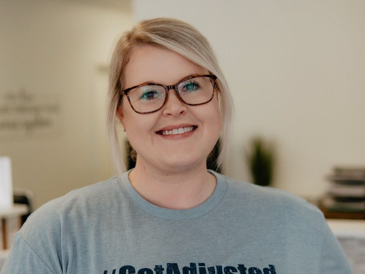 A woman wearing glasses and a t-shirt that says `` got adjusted '' is smiling for the camera.