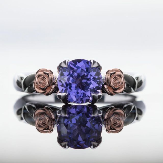 Silver ring with a purple gemstone flanked by two rose-shaped accents, mirrored on a glossy surface