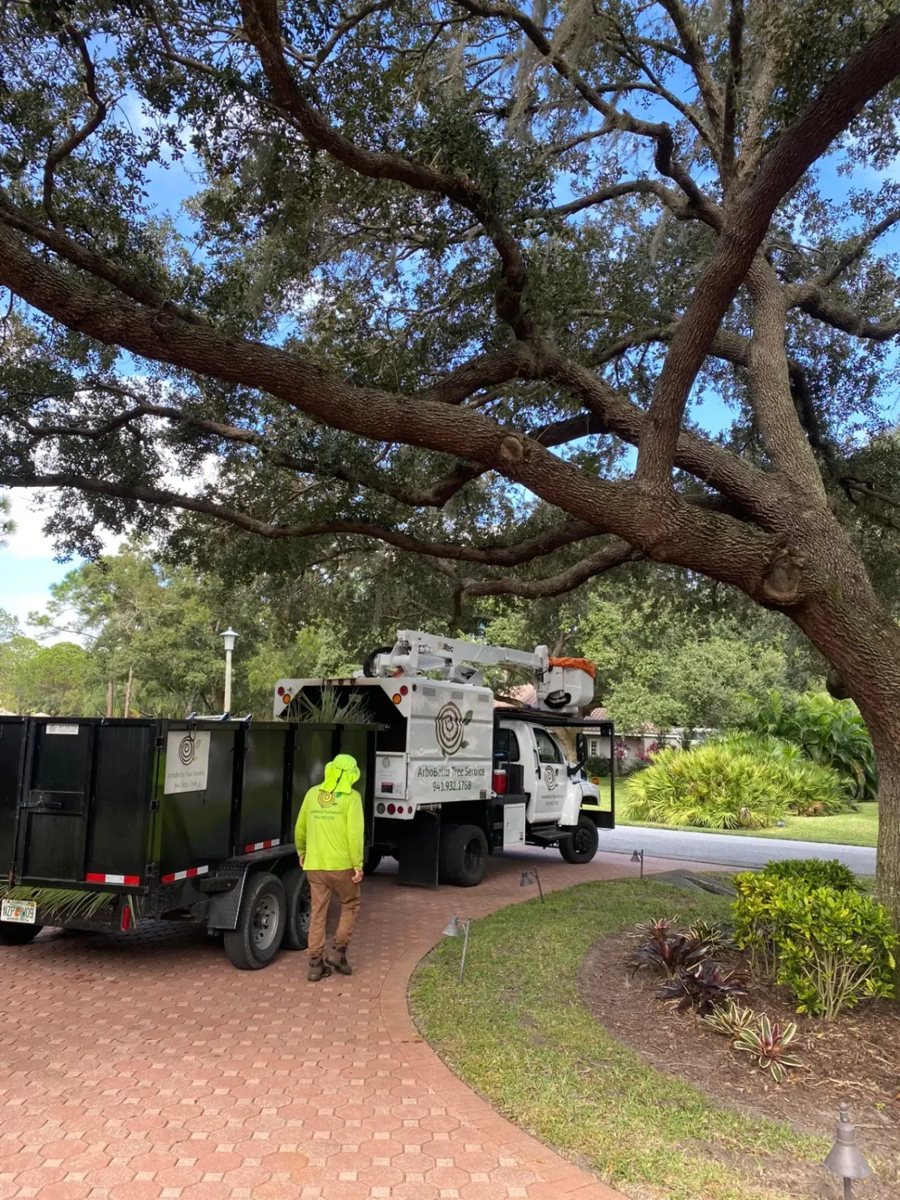 Worker in neon vest beside utility truck and trailer under a large tree on a curved brick driveway