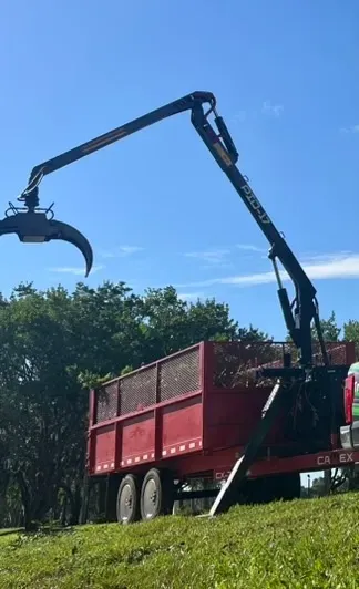 A red truck with a crane arm lifting a large claw over grass under a blue sky.