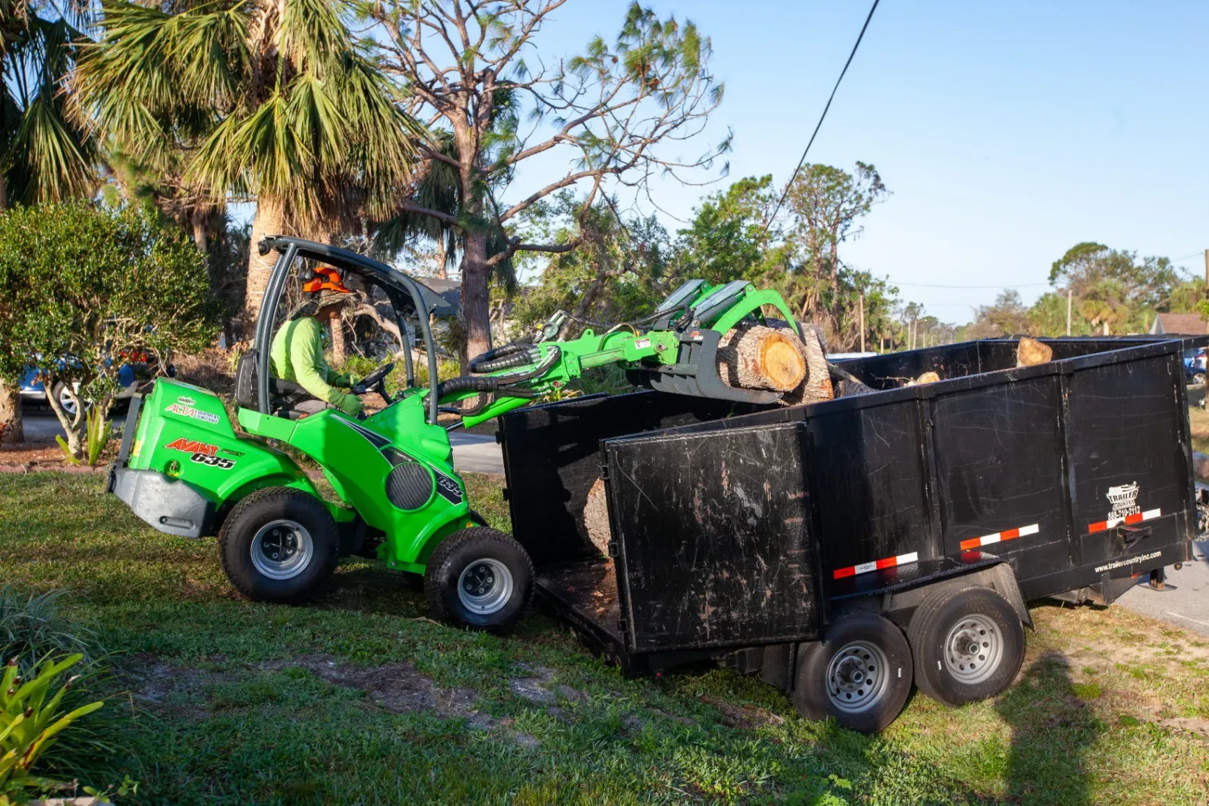 Green loader dumping debris into a black trailer outdoors near trees