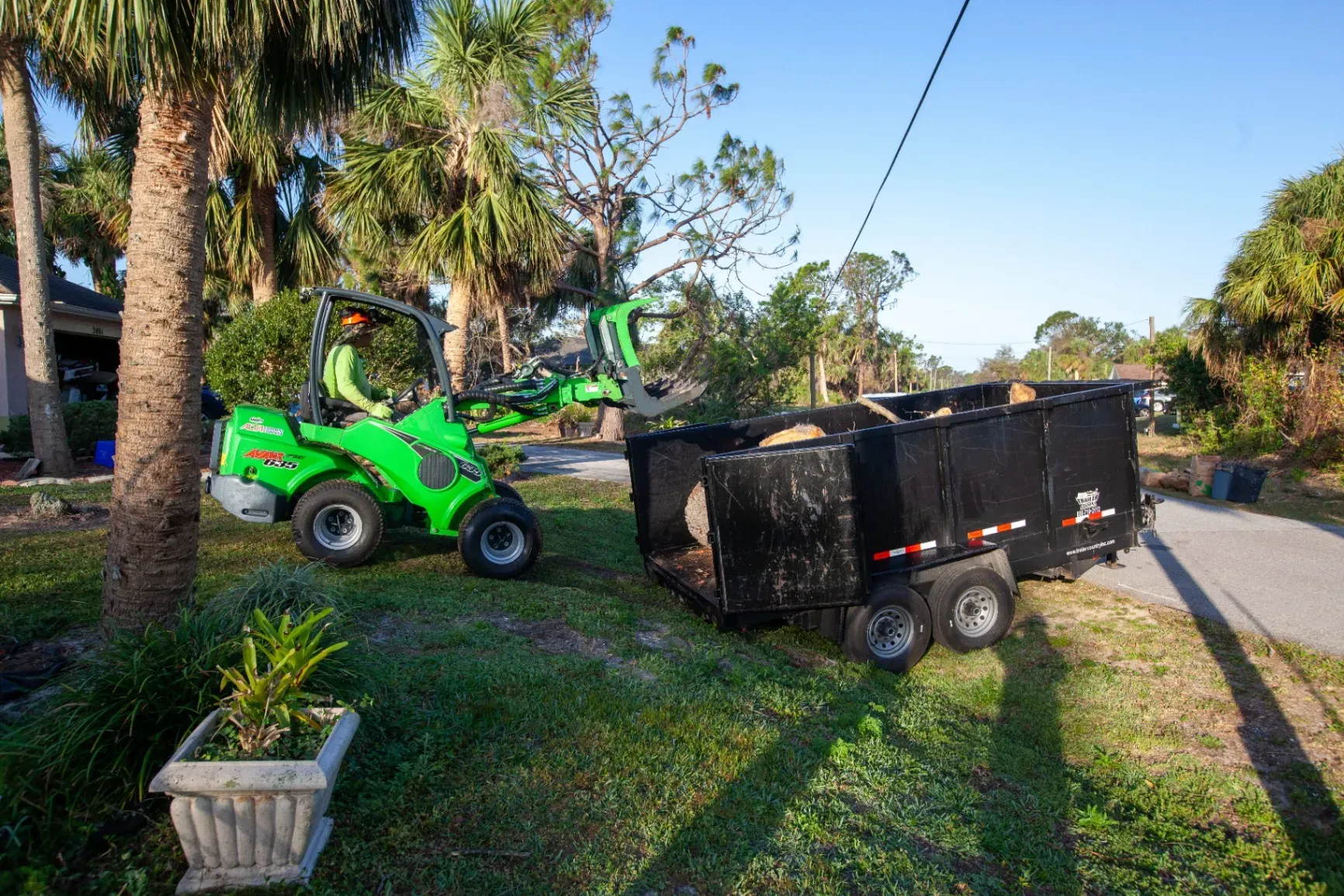 Green lawn tractor towing a black utility trailer on a grassy roadside with palm trees.