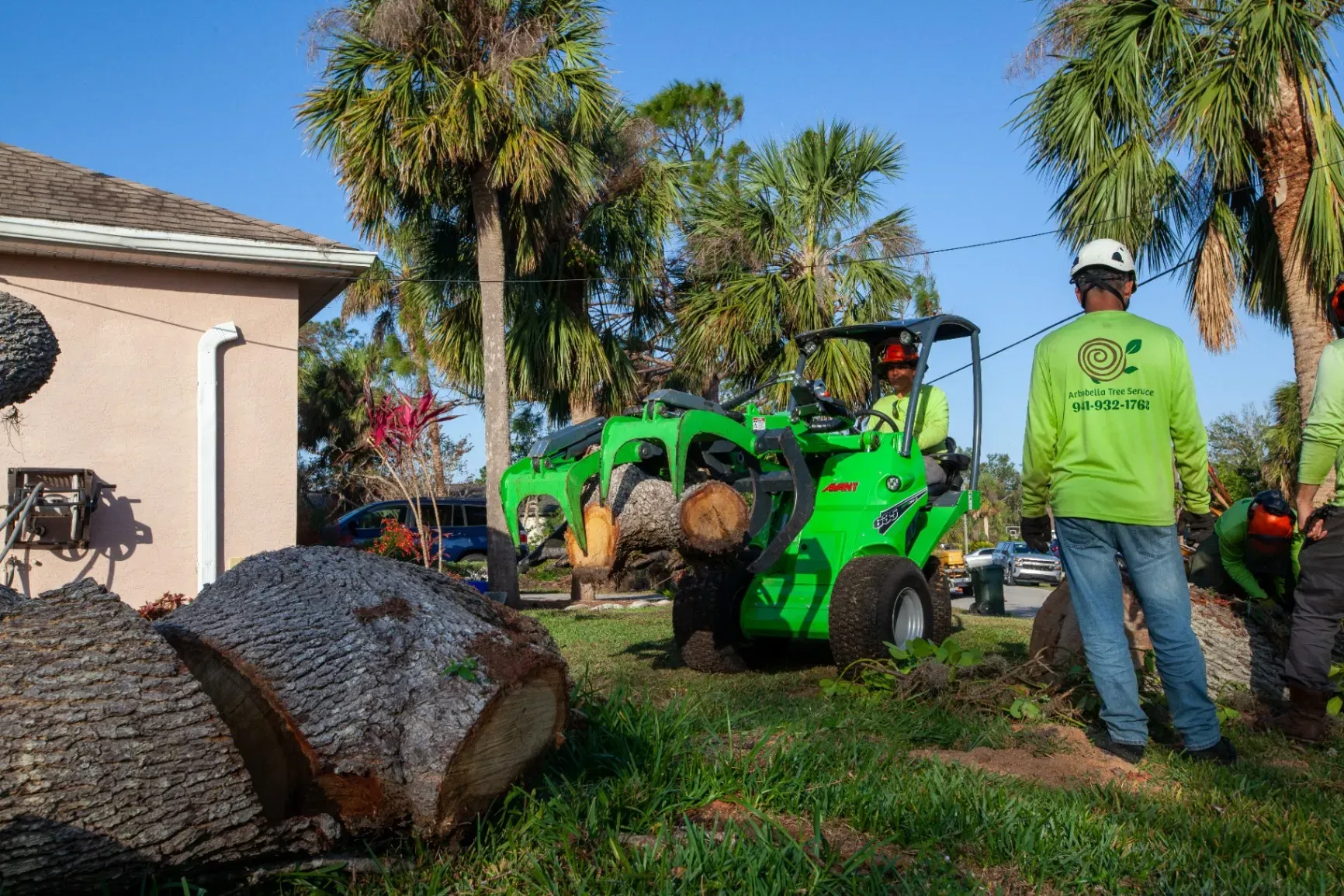 Landscapers with a green skid steer moving a large boulder in a sunny yard with palm trees