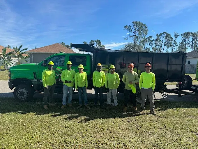 Seven workers in neon safety shirts posing beside a green truck and trailer on a grassy lot.