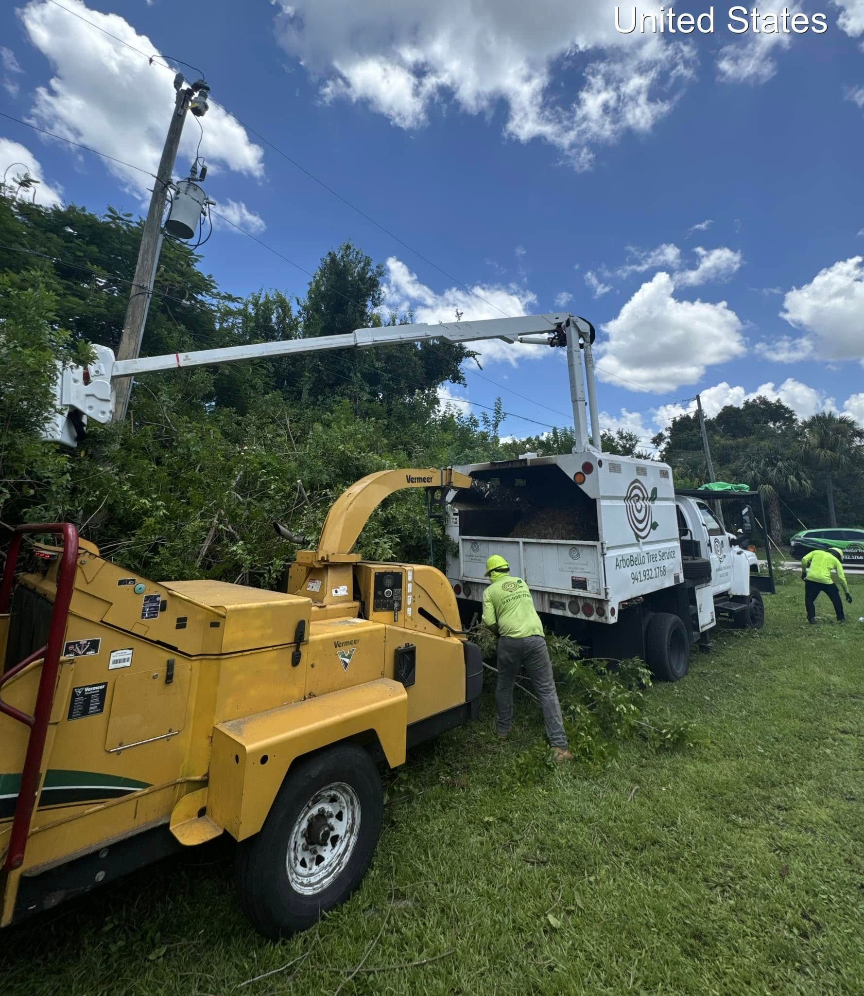 Utility crew using a yellow chipper beside a white truck in a grassy roadside area under a blue sky.