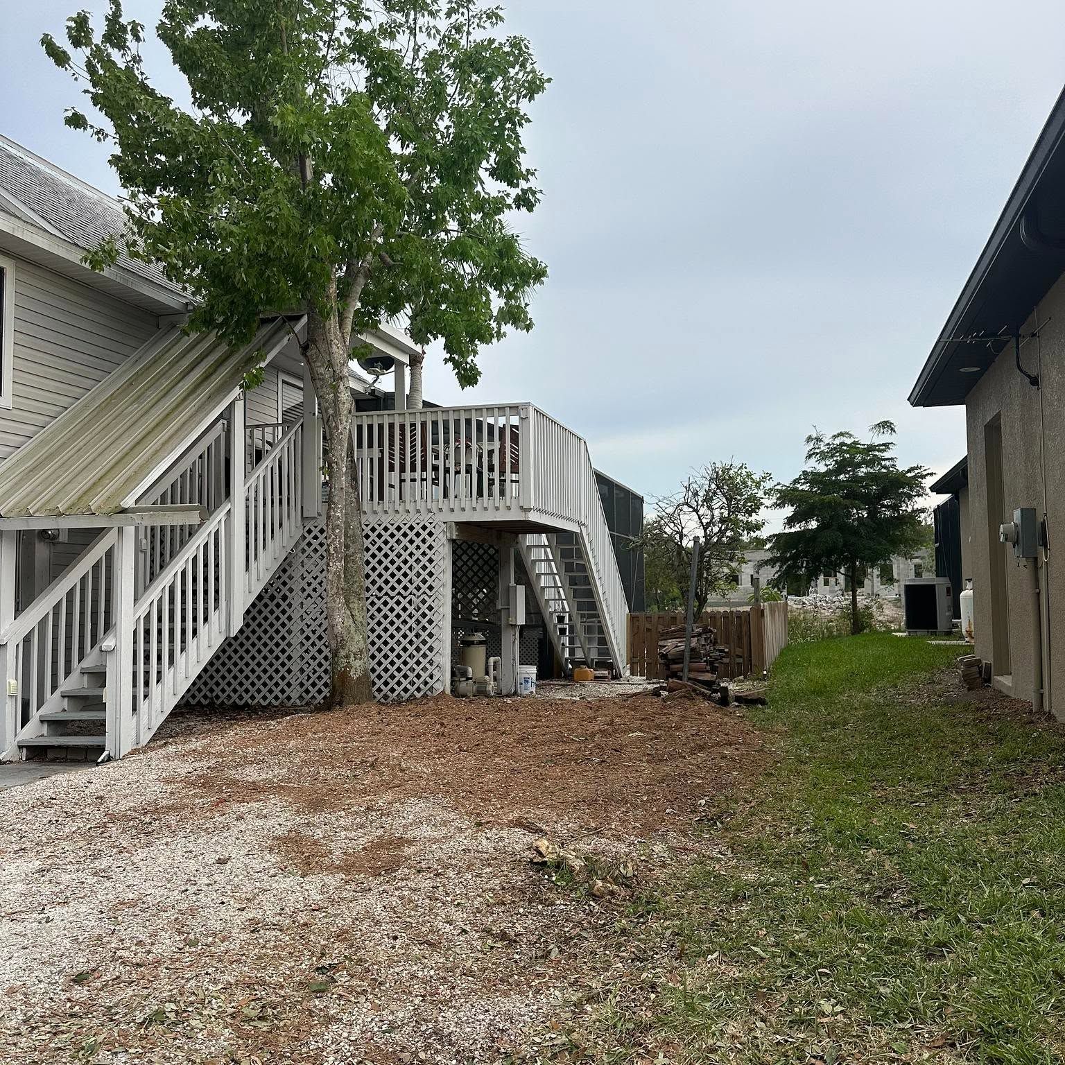 White-railed wooden deck beside a house, with stairs, a tree, and a gravel yard under cloudy sky