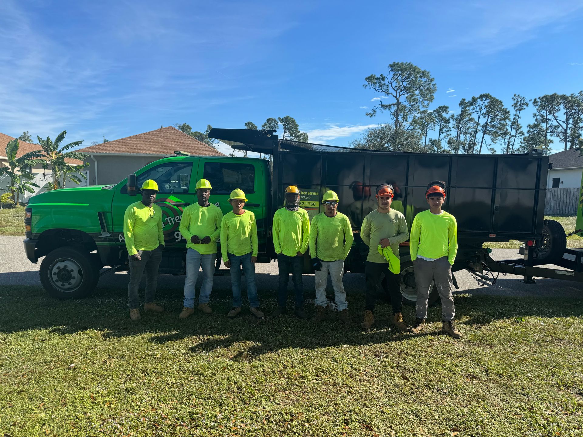 Seven workers in neon yellow shirts stand beside a green truck and trailer on a grassy lot.