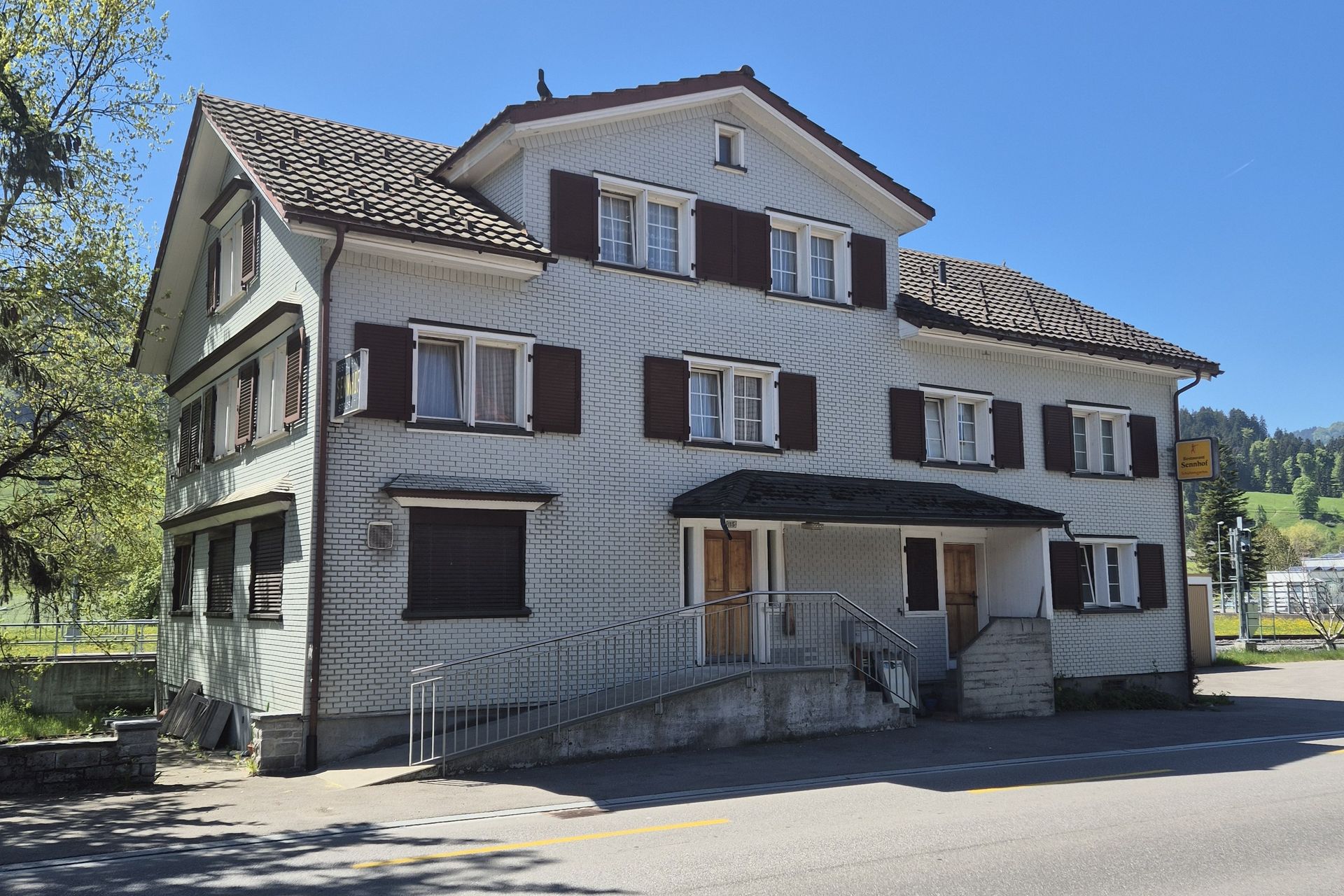 Ein zweistöckiges braunes Chalet mit weiß gerahmten Fenstern, einer überdachten Terrasse und einem Holzbalkon, eingebettet in einen Rasengarten.