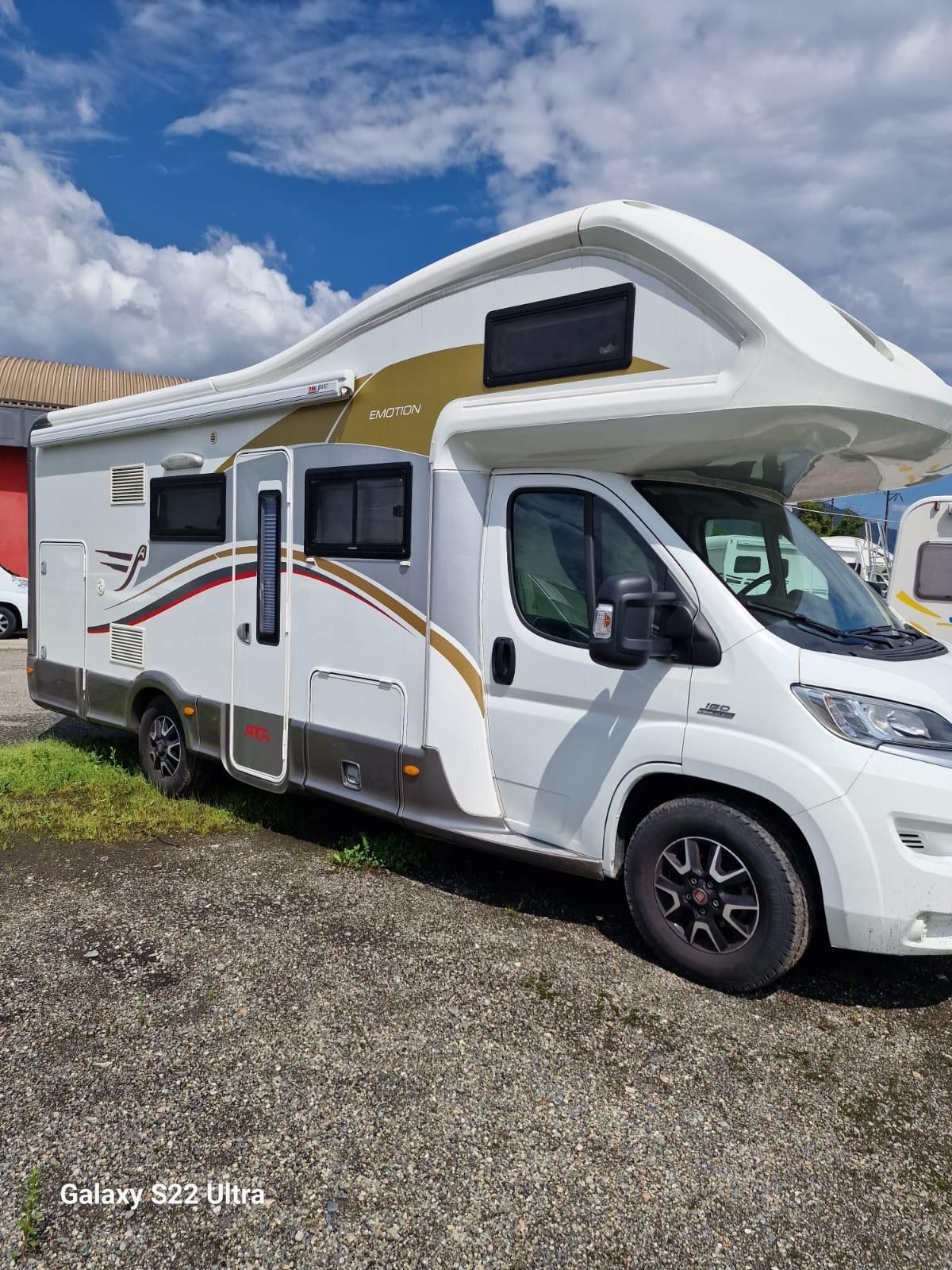 Camper bianco e oro parcheggiato all'aperto su un terreno ghiaioso contro un cielo azzurro.