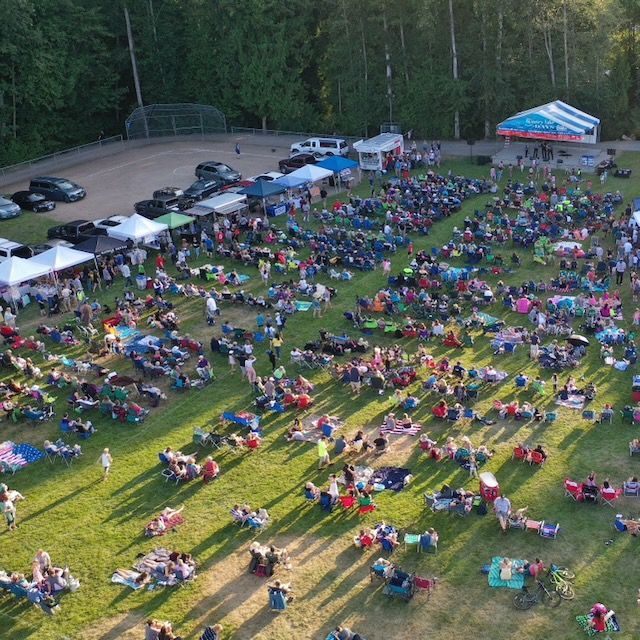 Large crowd at outdoor event on a grassy field, with a stage and tents in the background. Many people seated.
