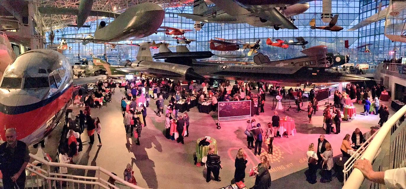 Large event inside a museum with aircraft hanging from the ceiling. People mingle, tables set.