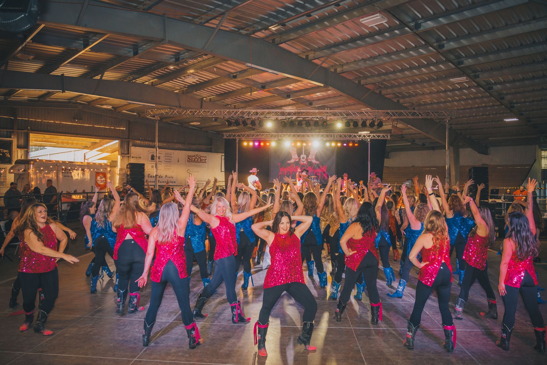 Large group of dancers in red tops and black pants, performing on a stage lit with spotlights.