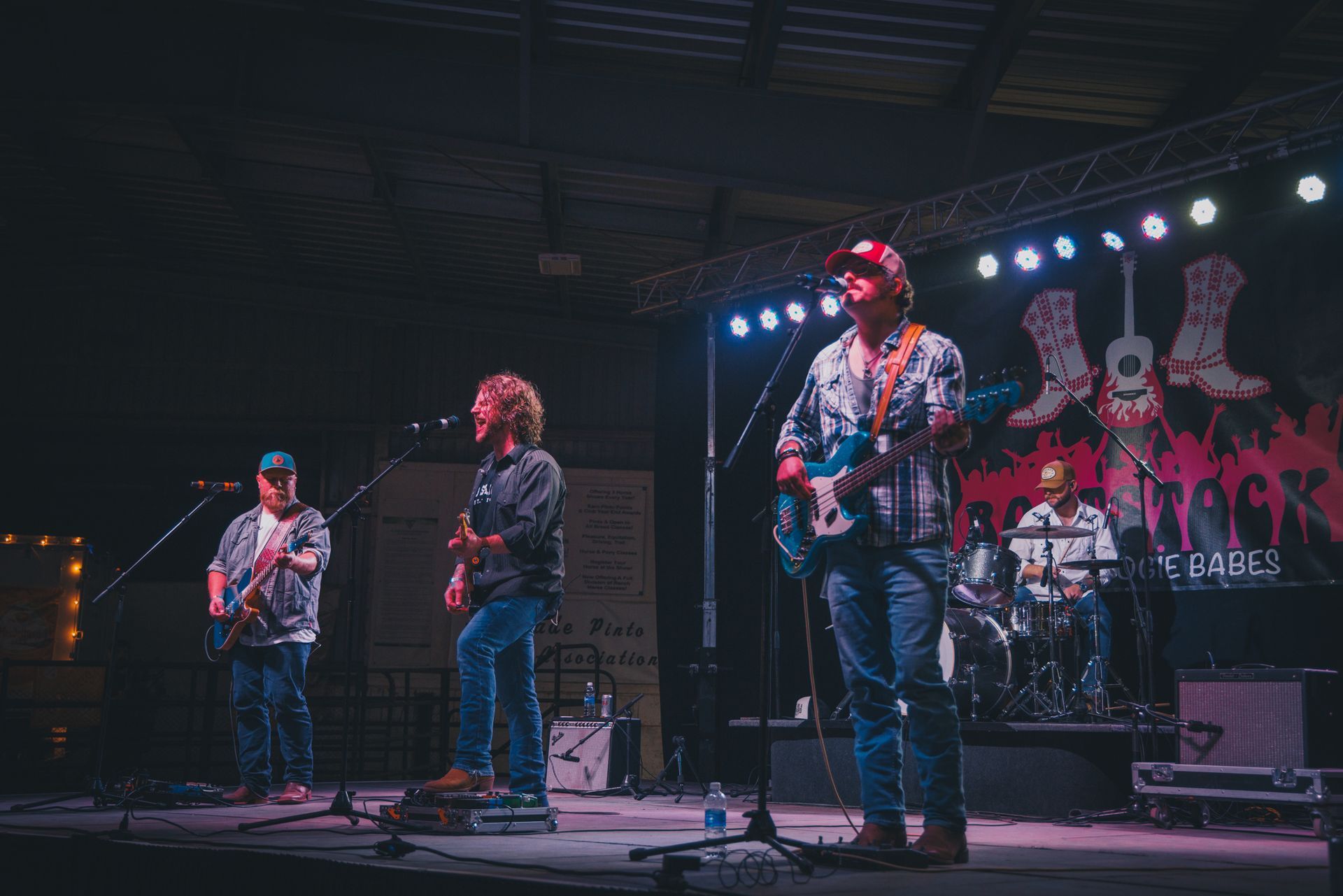 Band performing on stage at an outdoor event; musicians playing guitars, drums, and singing under stage lights.