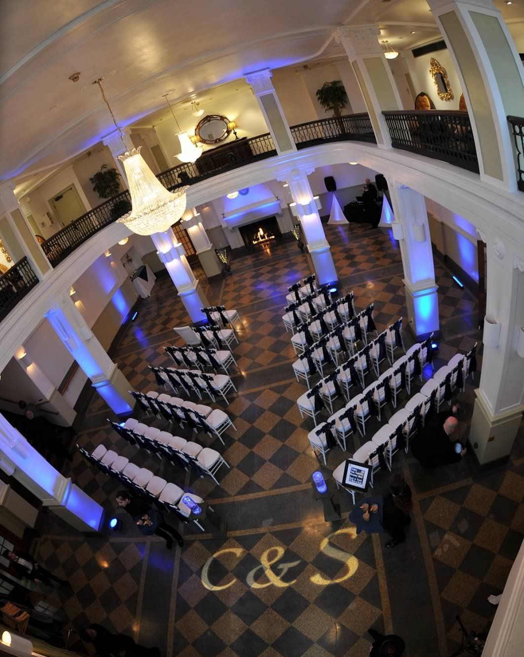 Ceremony venue, overhead view. Rows of chairs face a small stage. Blue lighting accents columns and initials on the floor.