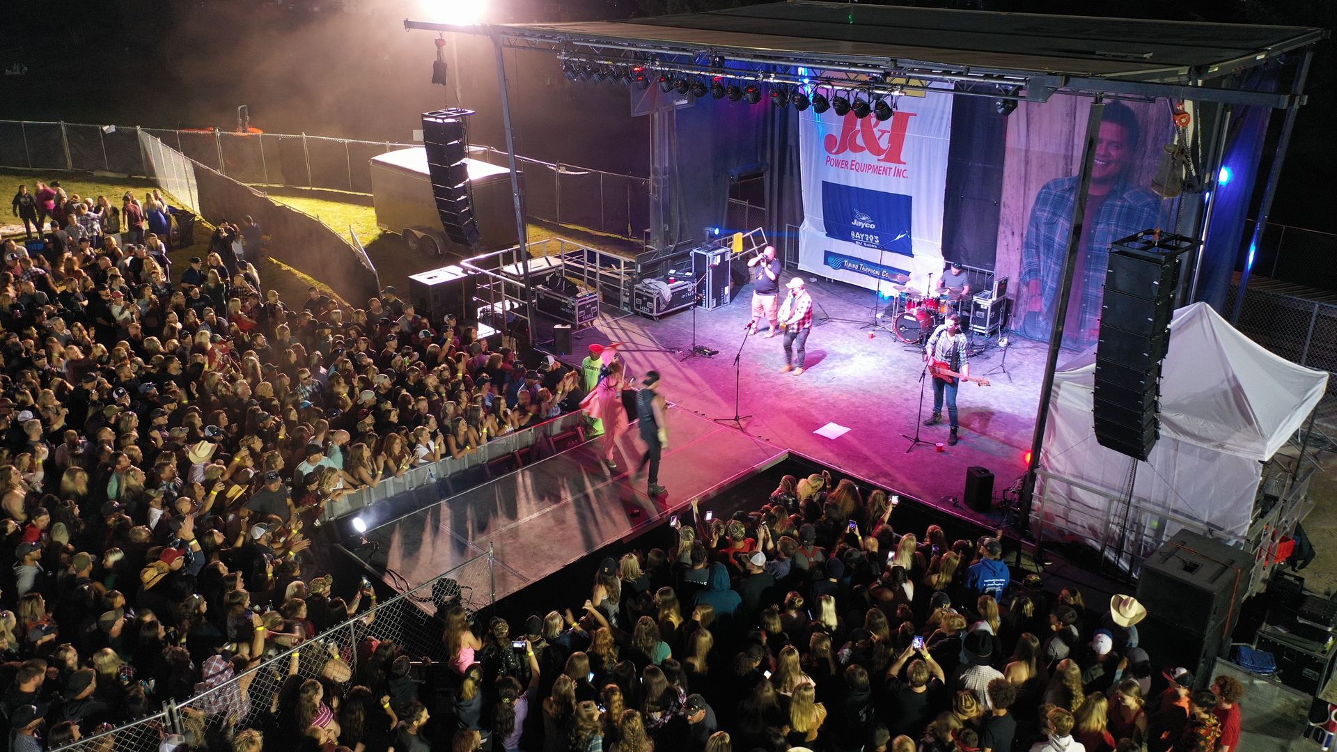 Band performs on outdoor stage for a crowd at night. Large backdrop. Lights shine down. People in the audience.