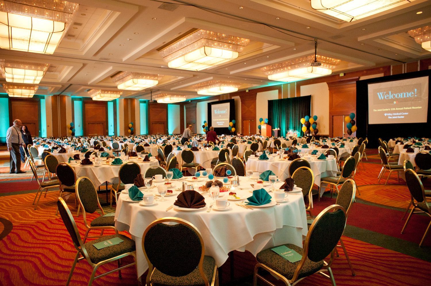 Large ballroom set for event; round tables with white linens, chairs, and centerpieces; screens and lighting.
