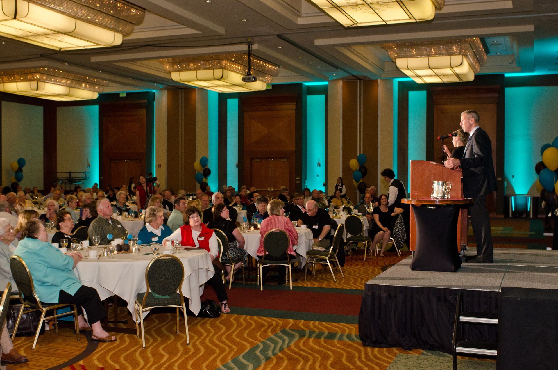 Man speaking at a podium on a stage in a large ballroom with tables and an audience.