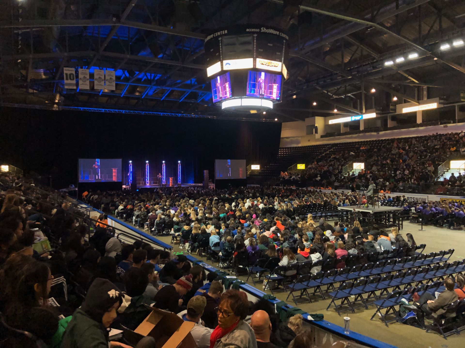 Large crowd at an indoor event, likely a conference. Stage with screens and lights. People seated, looking forward.
