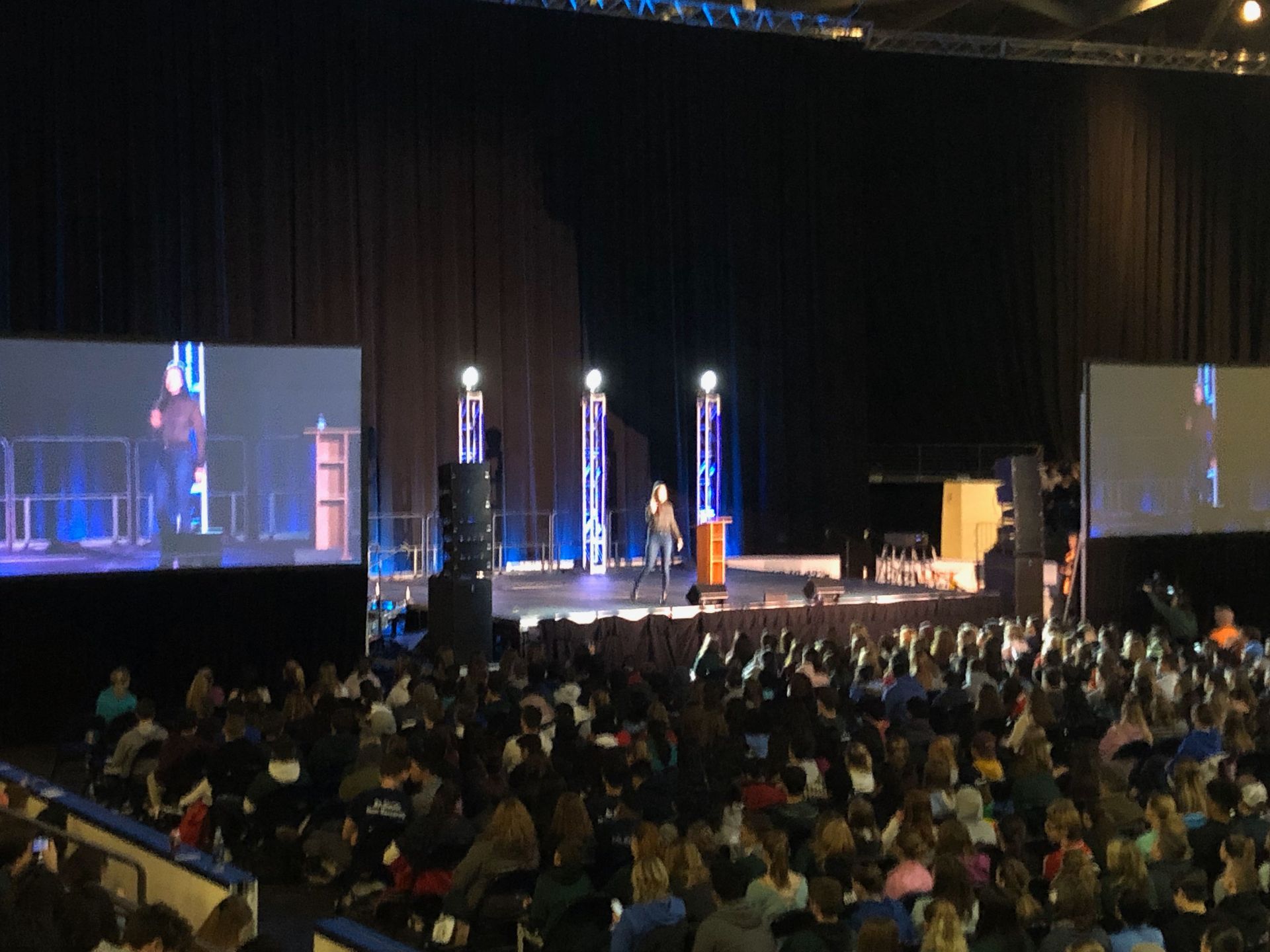Audience watches presenters on stage at a large indoor event with screens and lights.