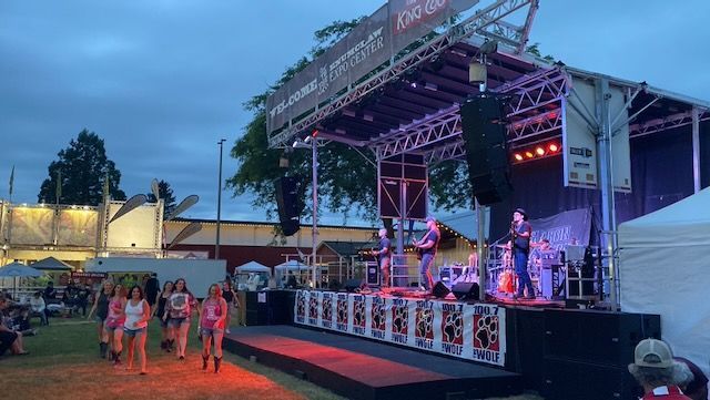 Band performs on outdoor stage at dusk, with crowd and fairground structures in background.