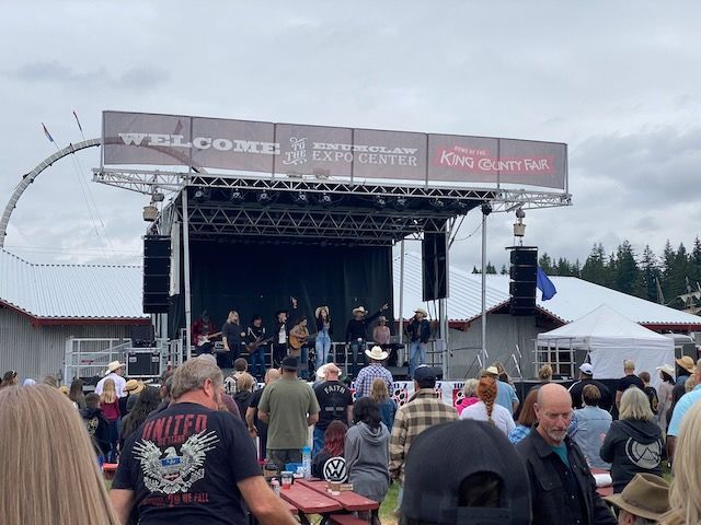 Live band performing on stage at the King County Fair. People watch from the crowd on a cloudy day.
