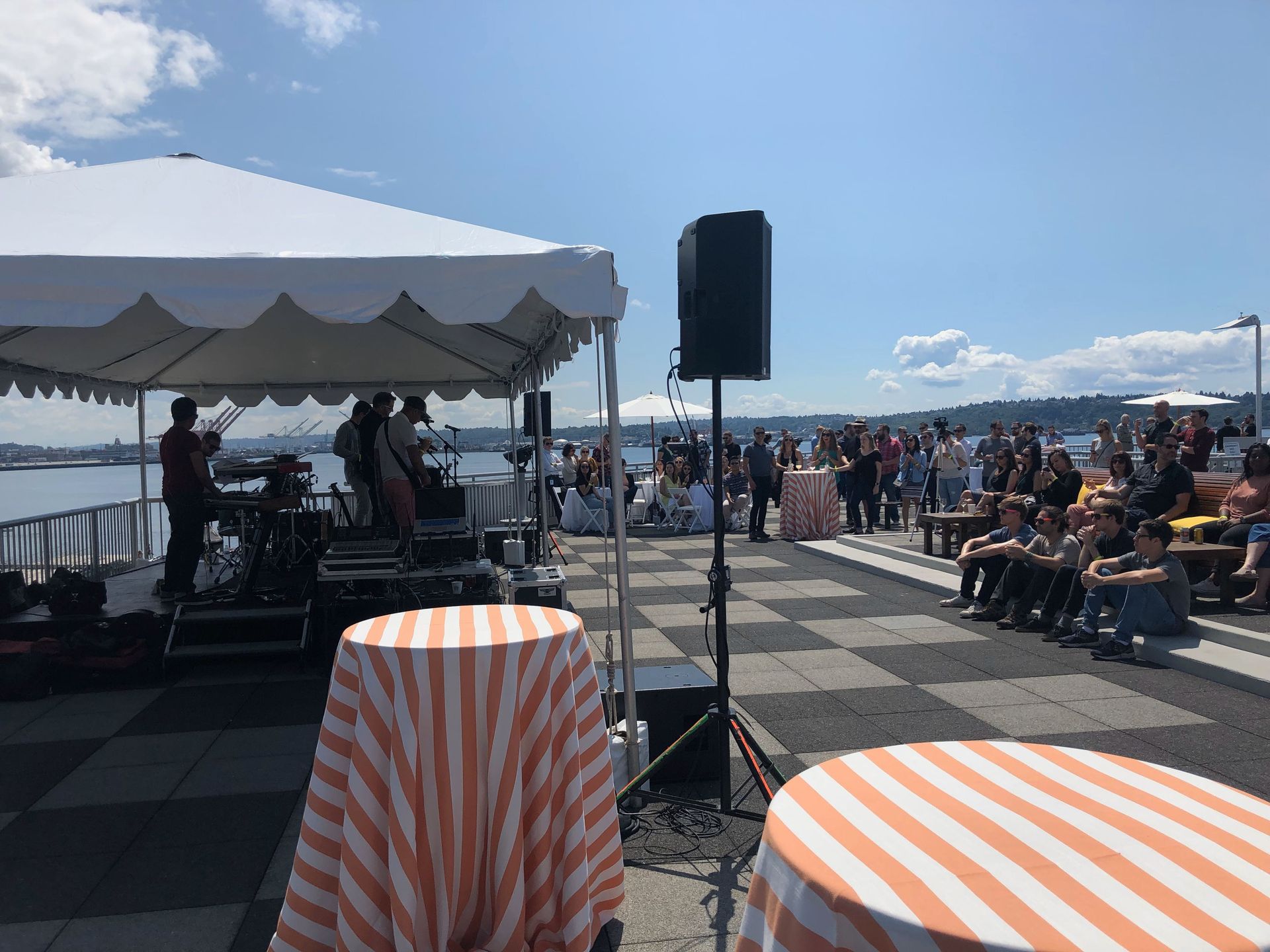 Band playing under a tent at an outdoor event near the water, with people in the audience.