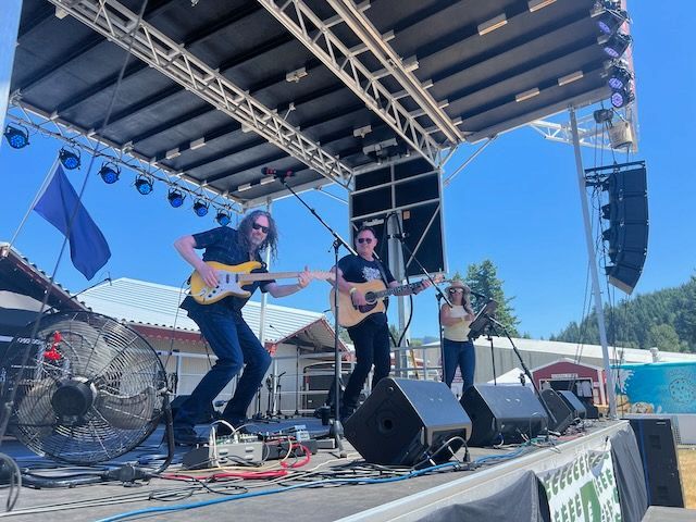 Band performing on outdoor stage, guitarist jumping, other musicians playing instruments, blue sky.