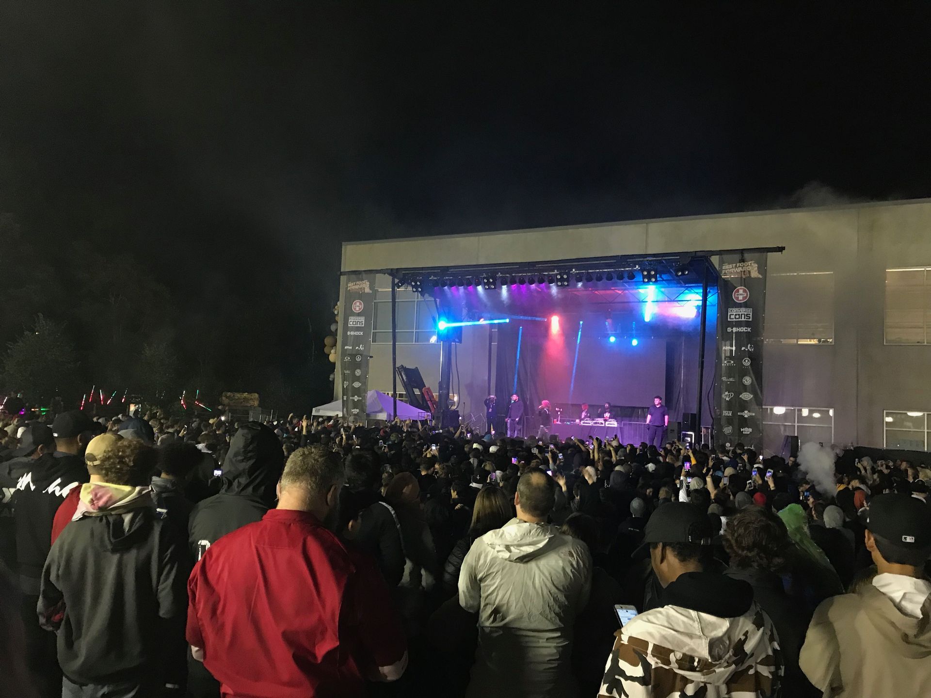 A large outdoor concert at night. Crowd faces illuminated stage with bright colorful lights.