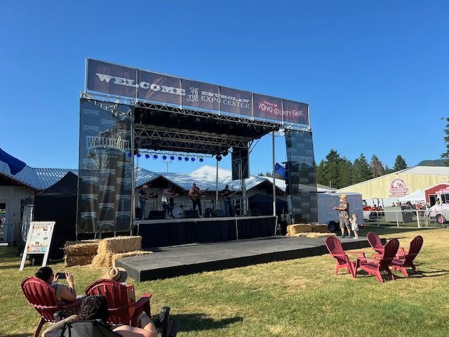 Stage set up for a concert in a grassy field. People in chairs face the stage. Blue sky.