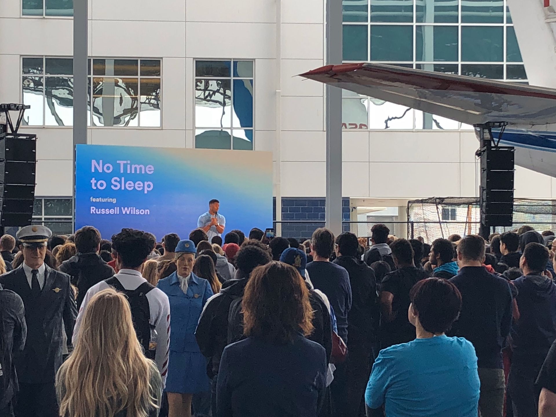 Man speaking at an event in a large hangar. Crowd watches; banner reads 