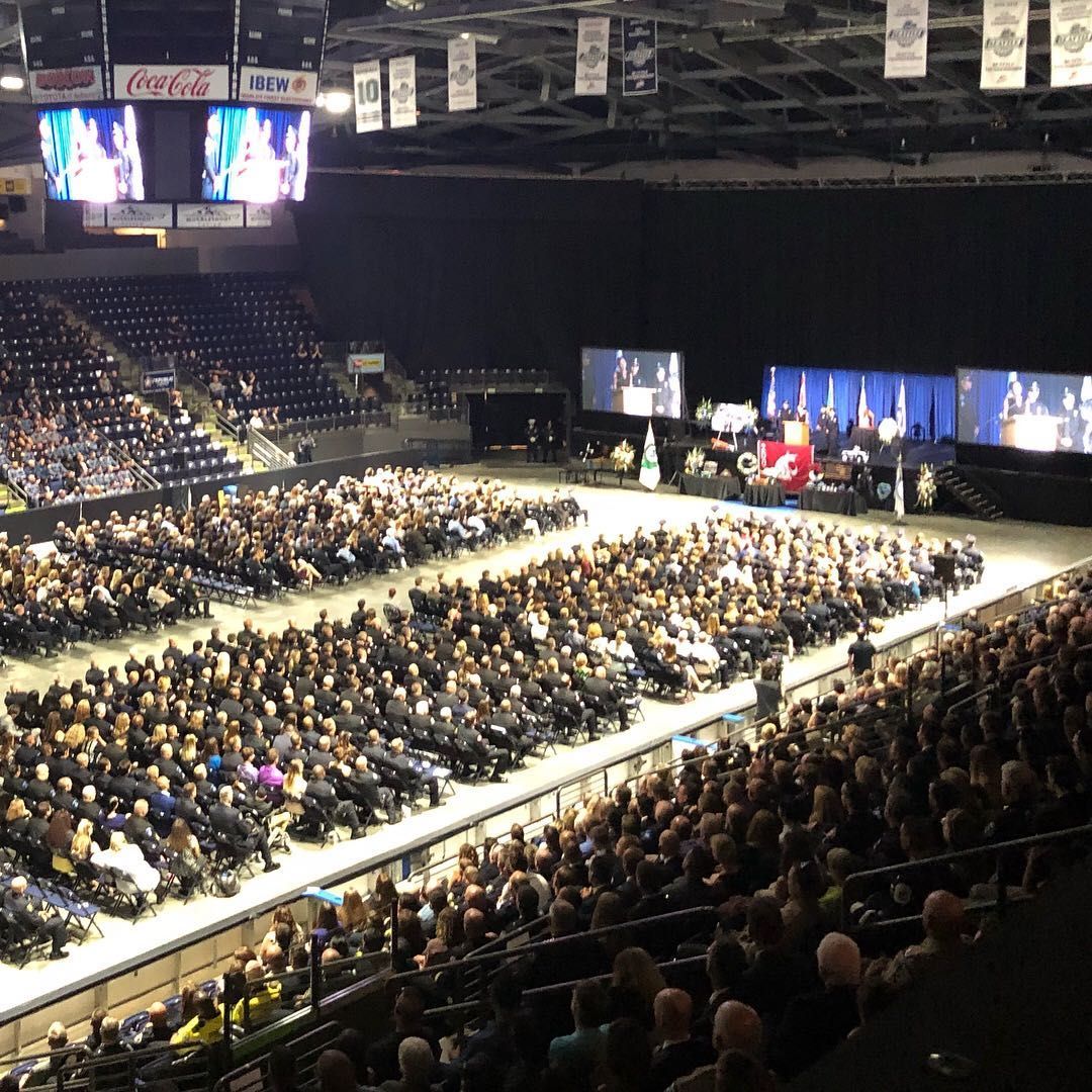 Large crowd in an arena for a graduation ceremony. Graduates in gowns fill the floor, audience in the seats.
